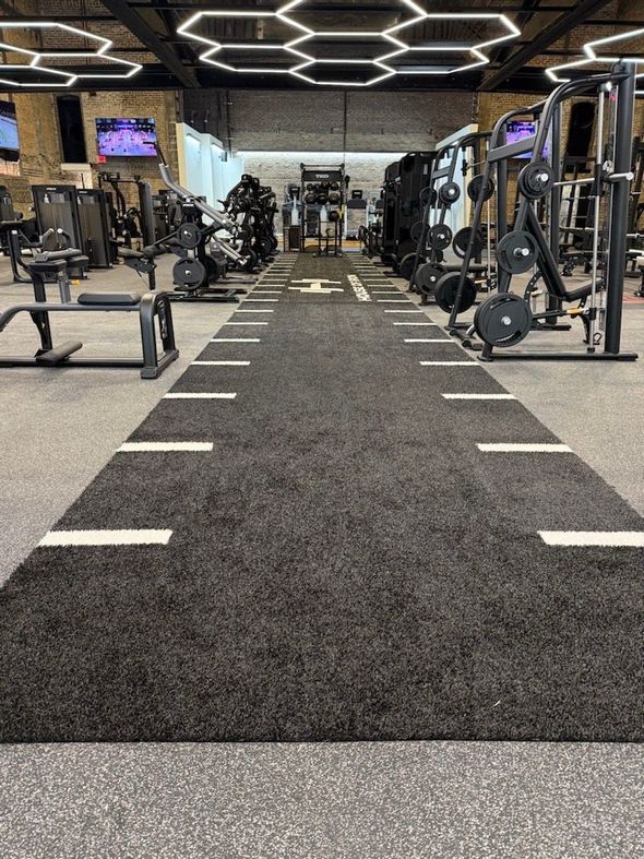 Modern gym interior with cardio and weight machines along a central turf lane and geometric ceiling lights
