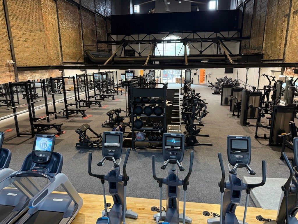Gym interior with rows of weight machines and stationary bikes on a turf floor, viewed from a cardio area