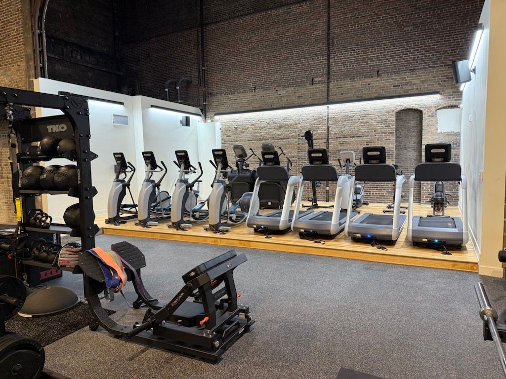 Compact gym with rows of cardio machines and weight benches on a carpeted floor, brick walls and exposed ceiling above