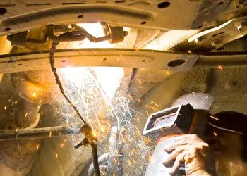 A man wearing a welding mask is welding under a car.