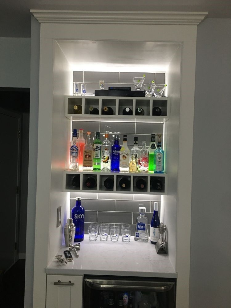 A built-in bar cabinet with white shelves, illuminated bottles, glassware, and a beverage fridge against a grey tile wall.