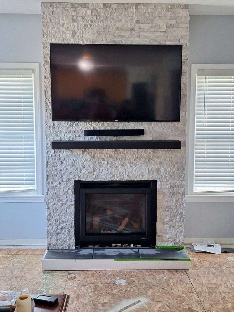 A stone-faced fireplace with a wall-mounted television and soundbar, set between two windows in a room under construction.
