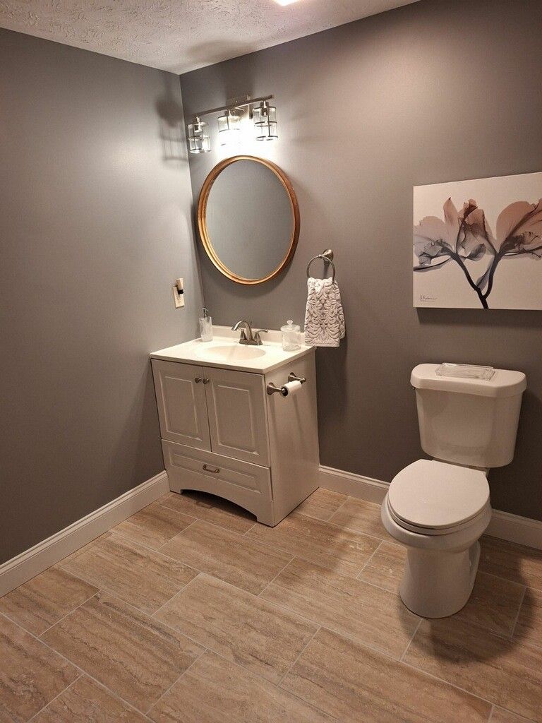 A modern bathroom with grey walls, a white vanity cabinet, a circular gold-framed mirror, and a white toilet.