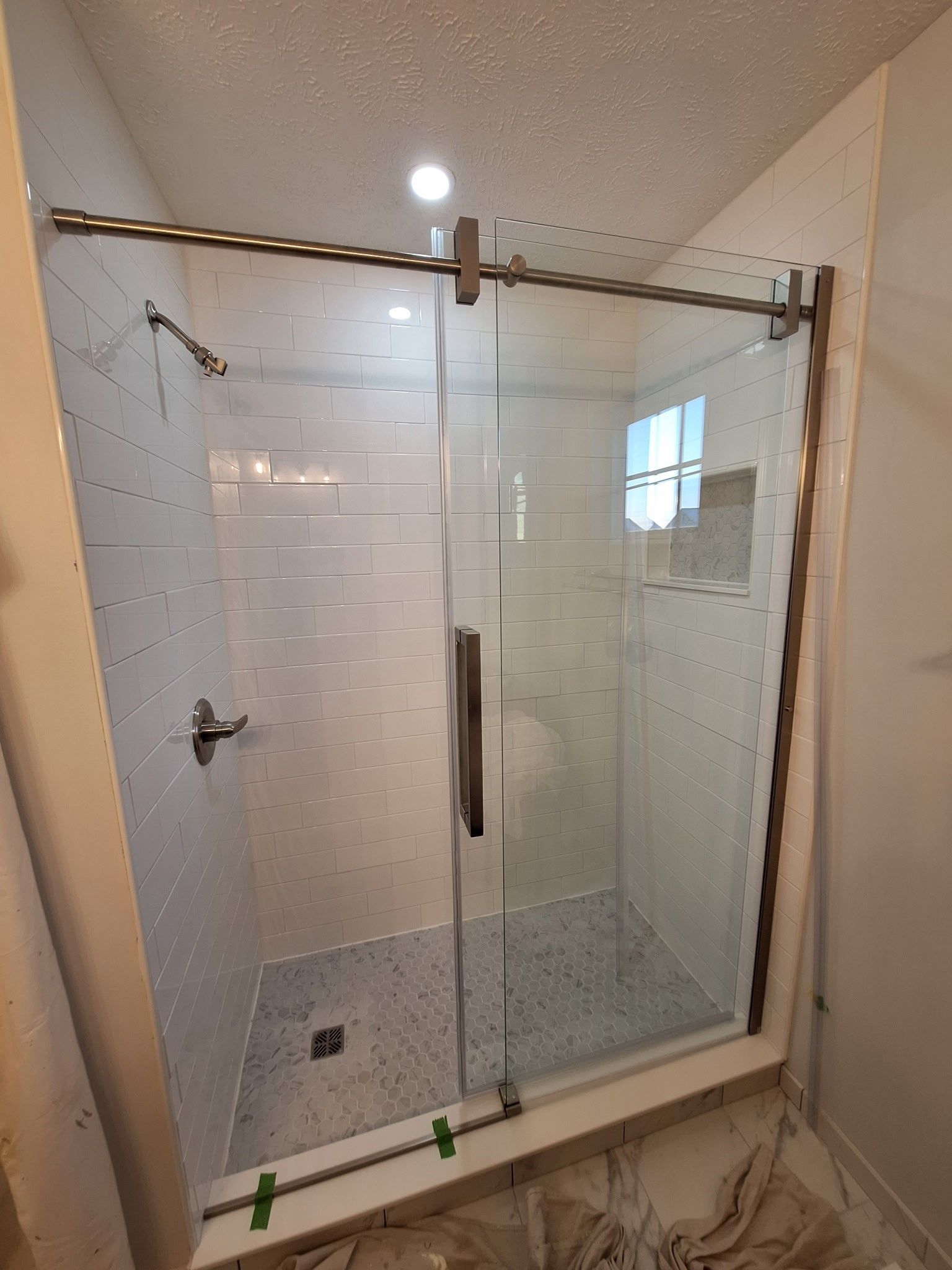 A glass sliding shower door installed in a bathroom with white tiled walls and a gray speckled floor.
