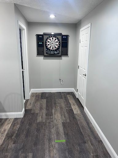 A dartboard mounted on a grey wall in a hallway with dark wood-look flooring and a green floor-taped throw line.