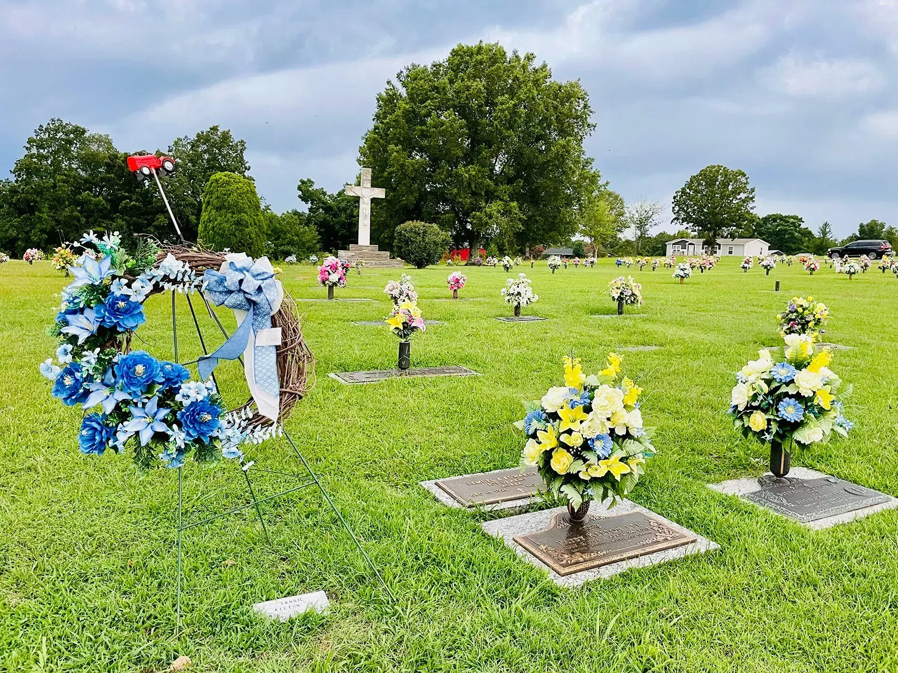 Floral wreaths and bouquets on graves in a sunny cemetery with a white cross in the background