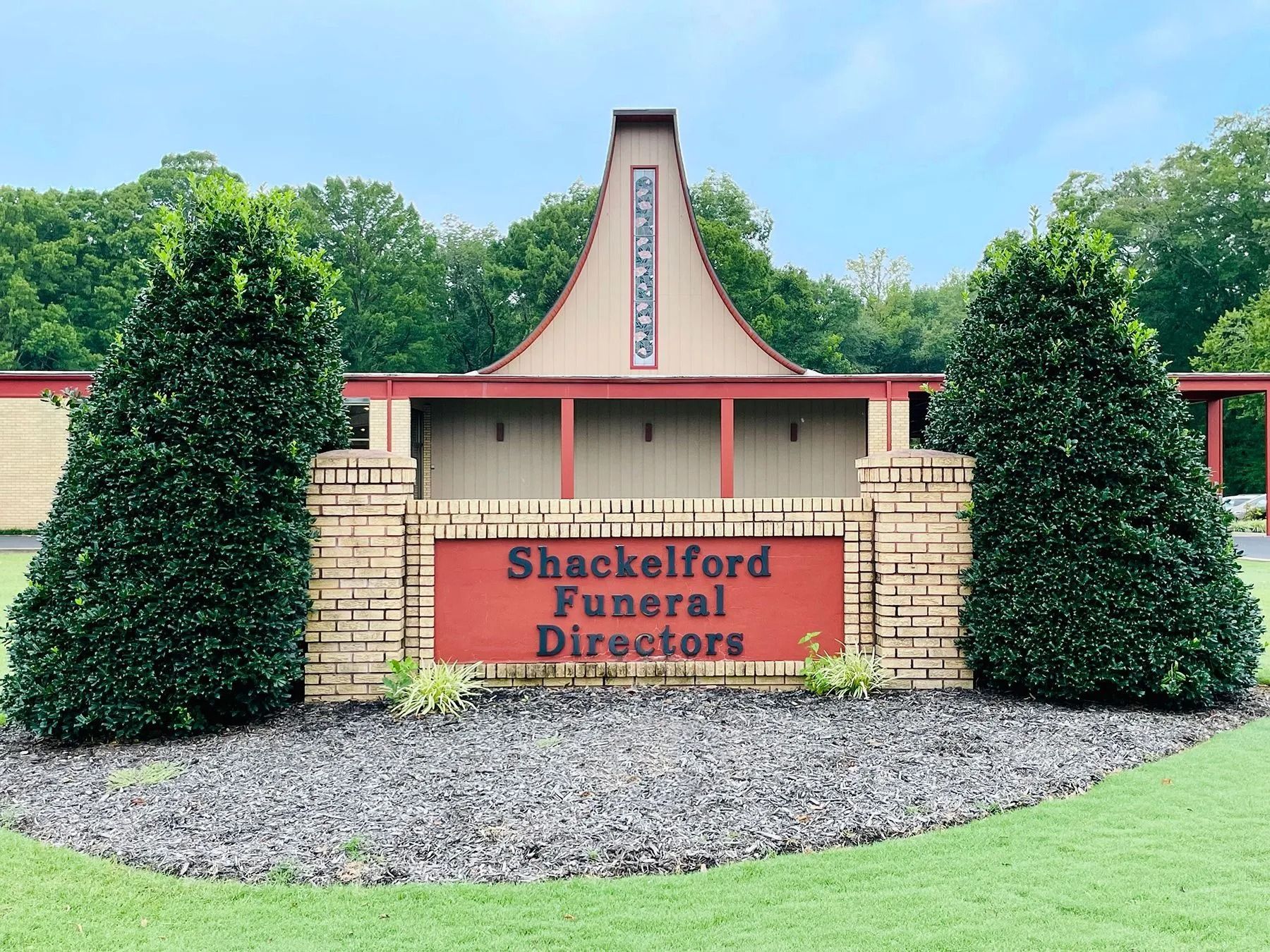 Shackelford Funeral Directors sign between trimmed evergreens and a brick building facade