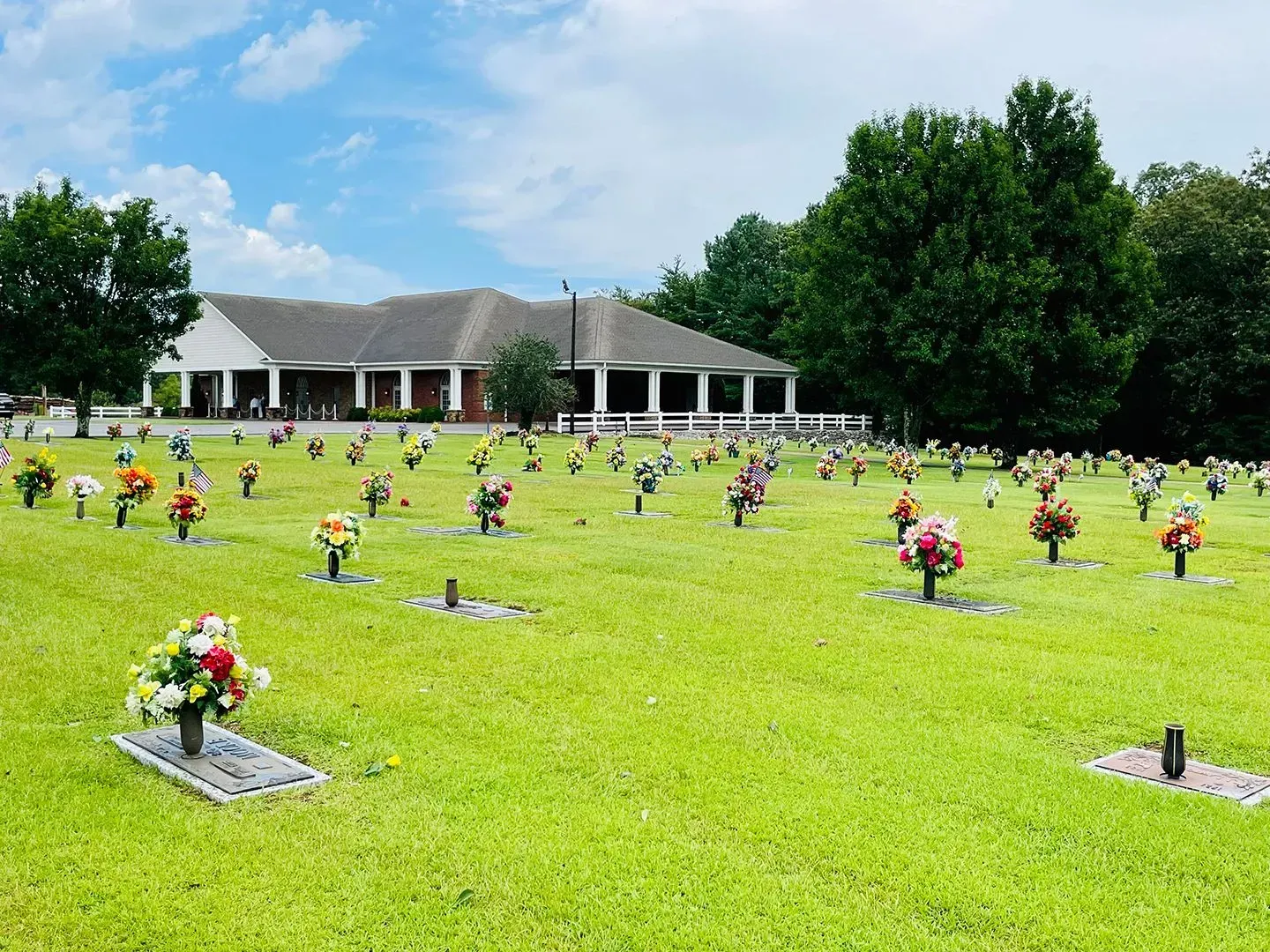 Grassy cemetery with many flower arrangements and a white memorial building under a cloudy sky
