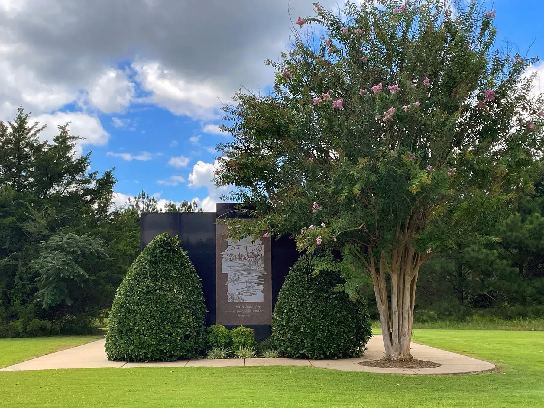 Park sign flanked by shrubs and a tree beside a walkway under a partly cloudy blue sky