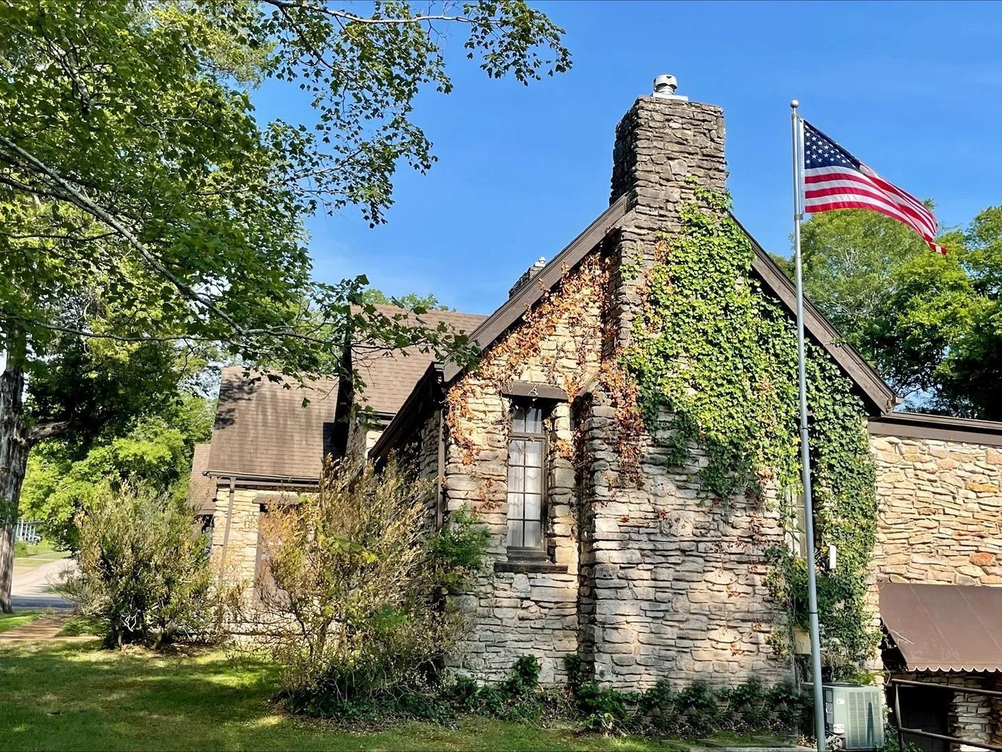 Stone cottage with a chimney and an American flag flying beside it under leafy trees.