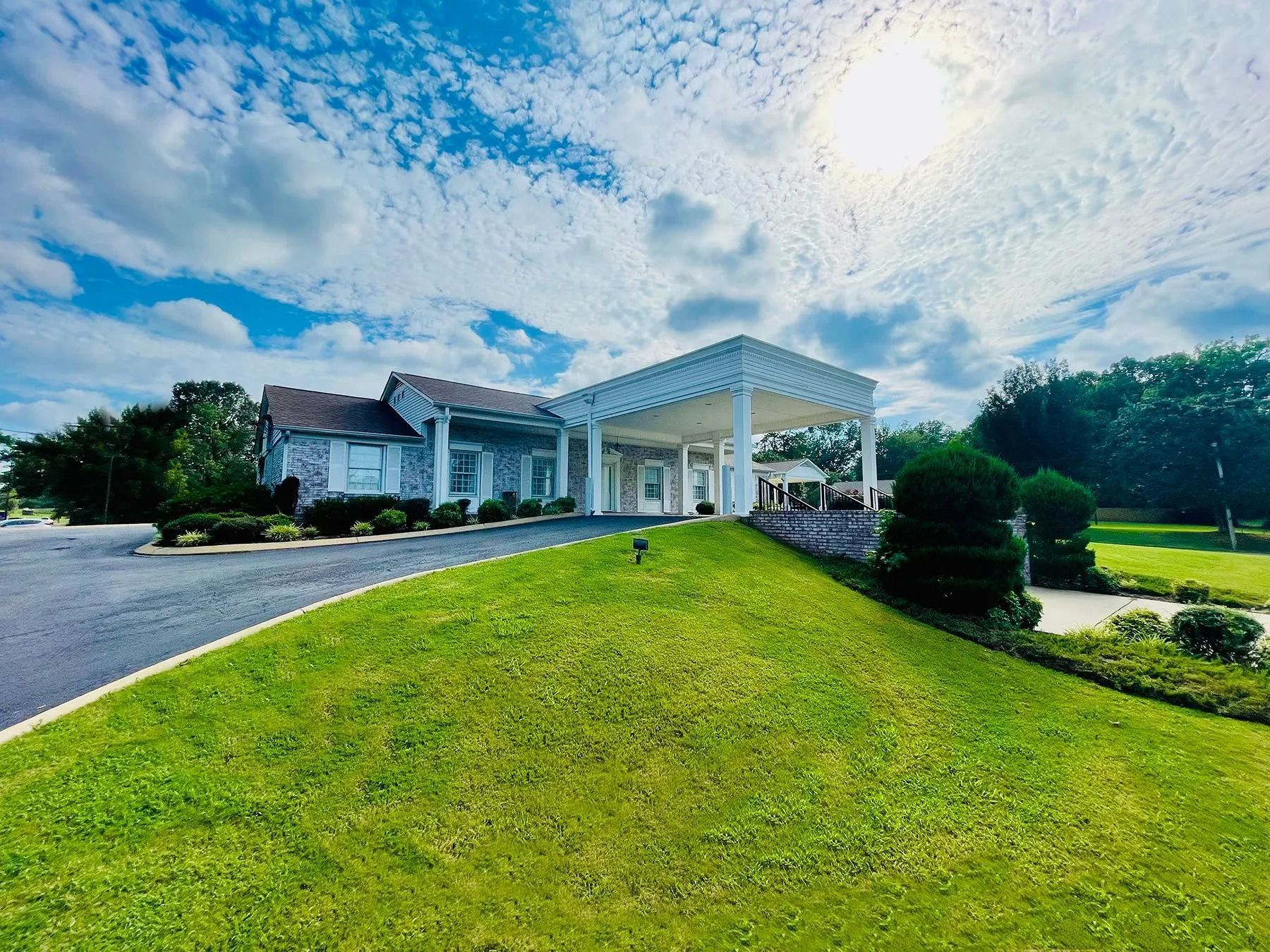 Large white house with a covered driveway, set behind a bright green lawn under a cloudy blue sky
