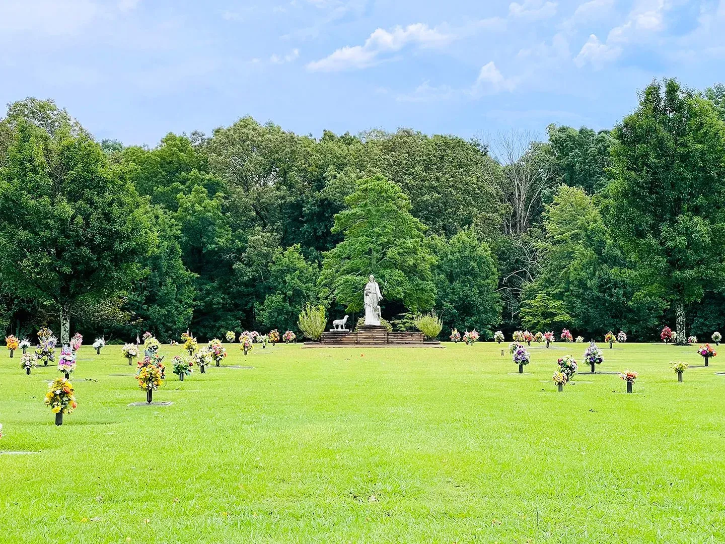 People scattered across a bright green lawn in a park, with trees and a small statue in the background.