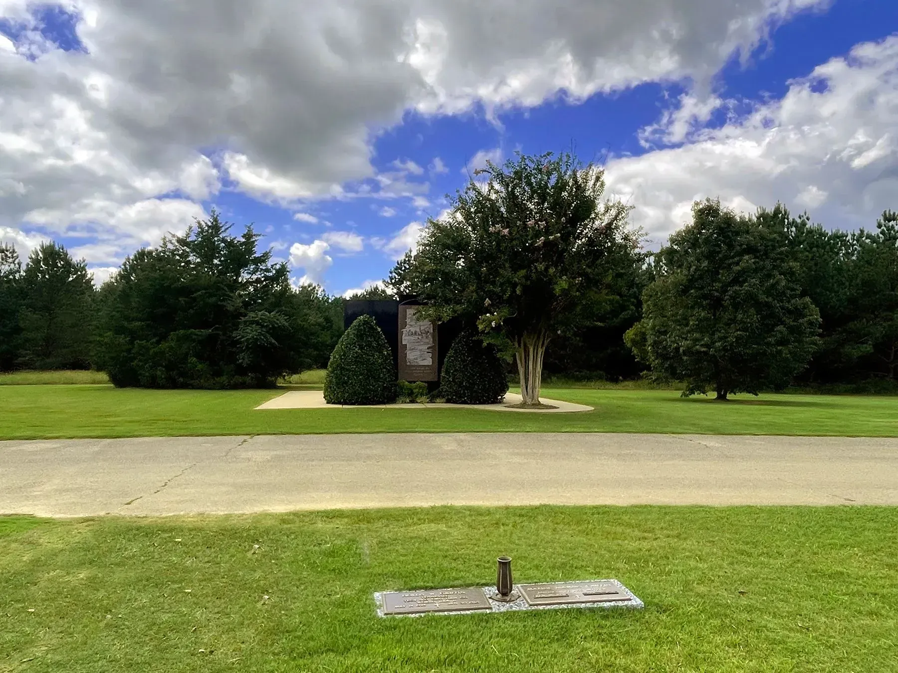 Grassy roadside park with trees, a small monument, and blue sky with clouds