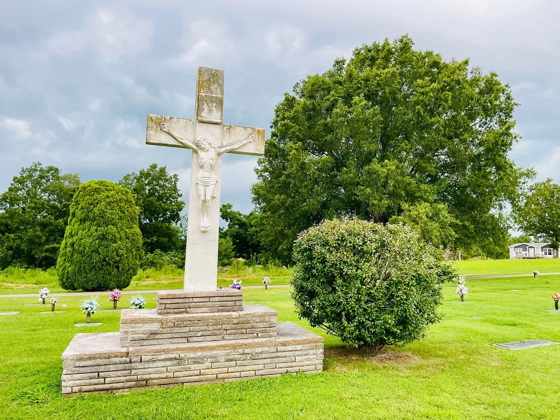 Stone cross monument in a green cemetery with trees and cloudy sky
