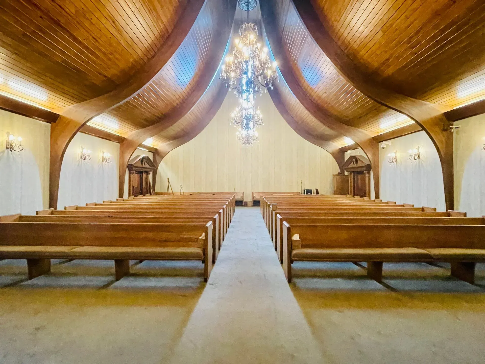 Empty church interior with wooden pews, a central aisle, and vaulted wood ceiling with chandeliers