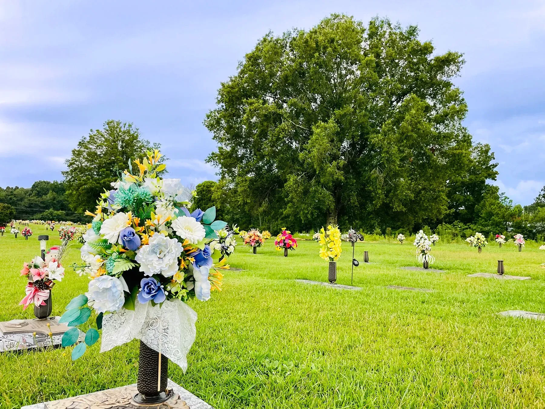 Flower-covered graves in a green cemetery under a large tree on a cloudy day
