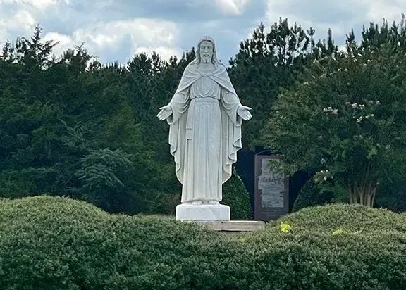 White statue of Jesus standing in a landscaped garden with trees and cloudy sky behind it