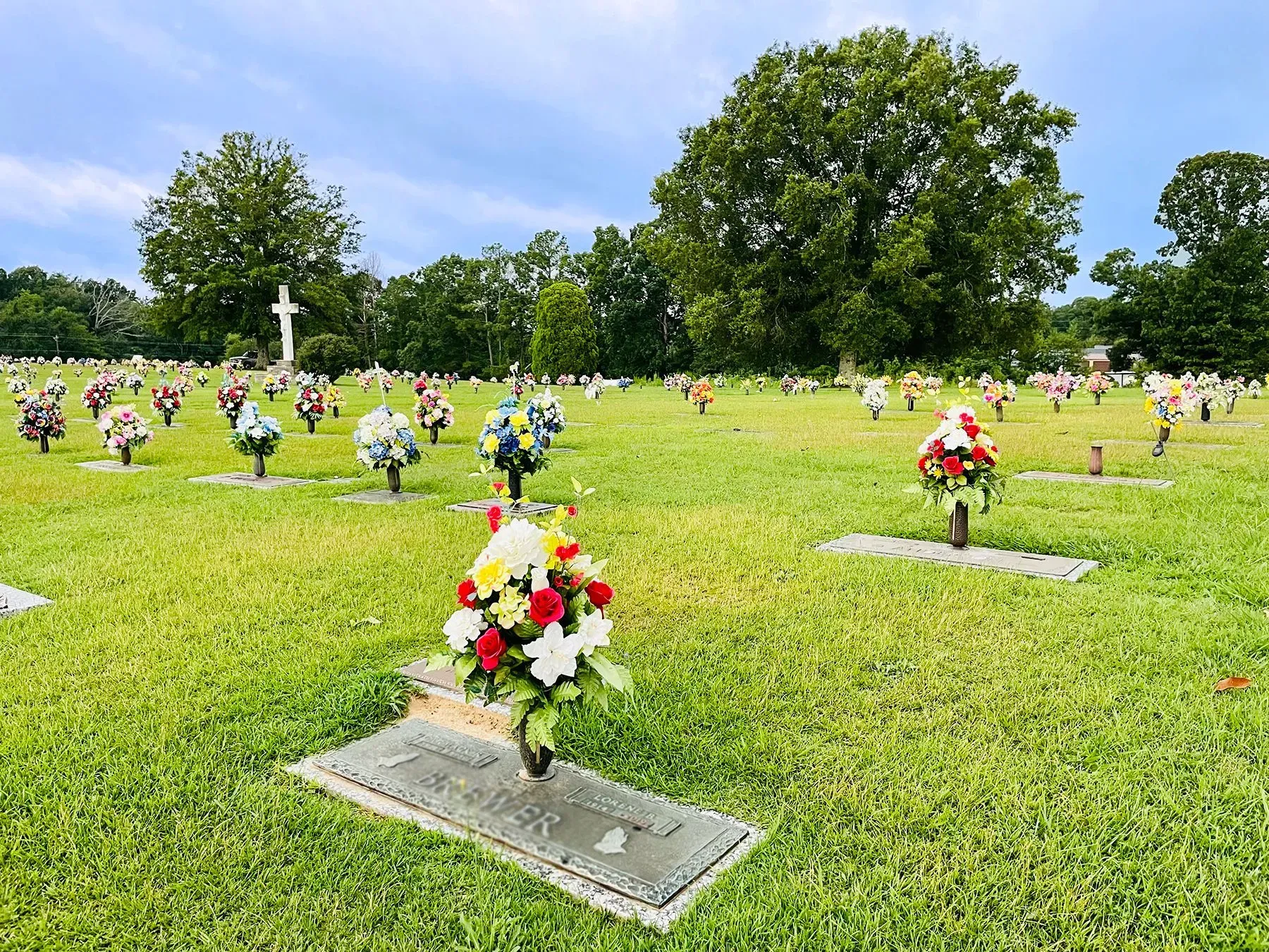 Cemetery with rows of flower-covered graves on a green lawn under a partly cloudy blue sky