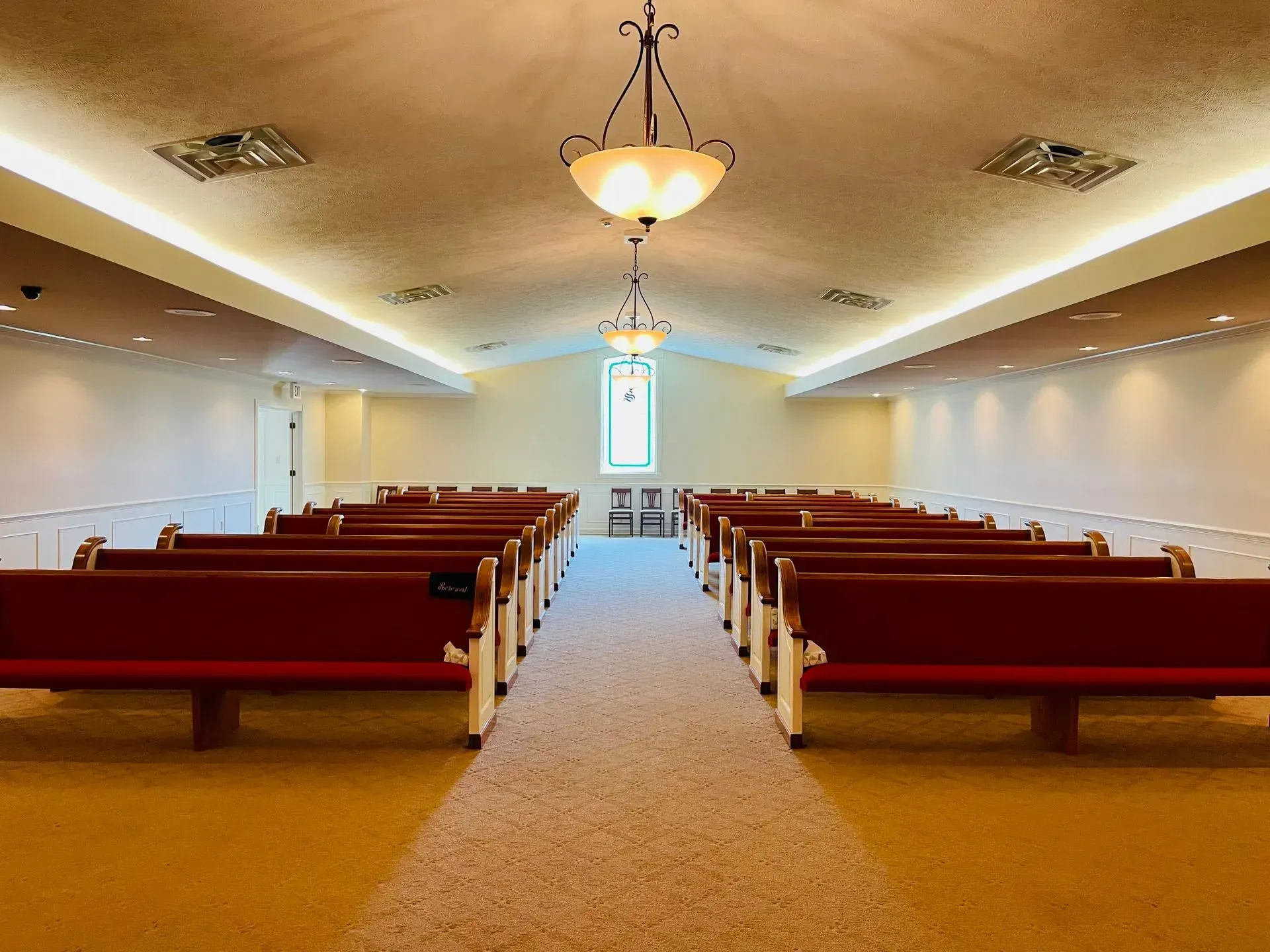 Empty church sanctuary with red pews, beige carpet, and a central chandelier facing a bright front window