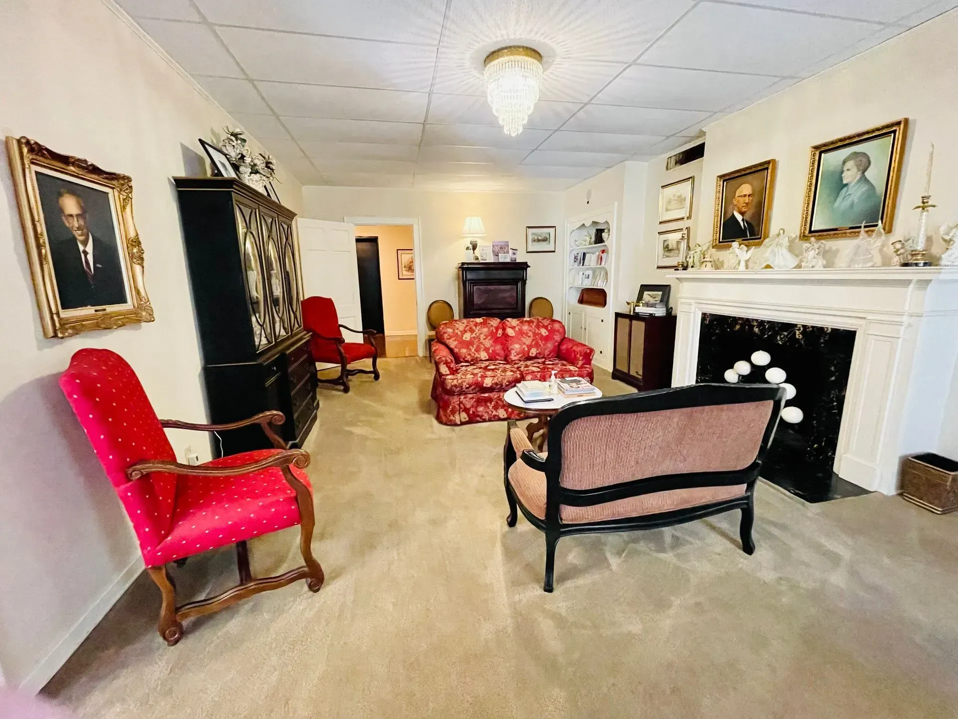 Bright living room with red and black vintage chairs, floral sofa, and a white fireplace with framed portraits.