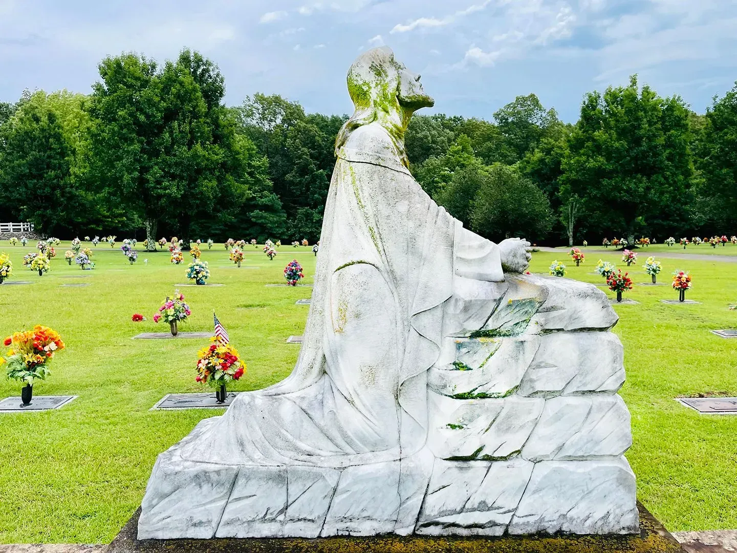 Marble statue of a cloaked figure in a green cemetery with colorful flower tributes and trees