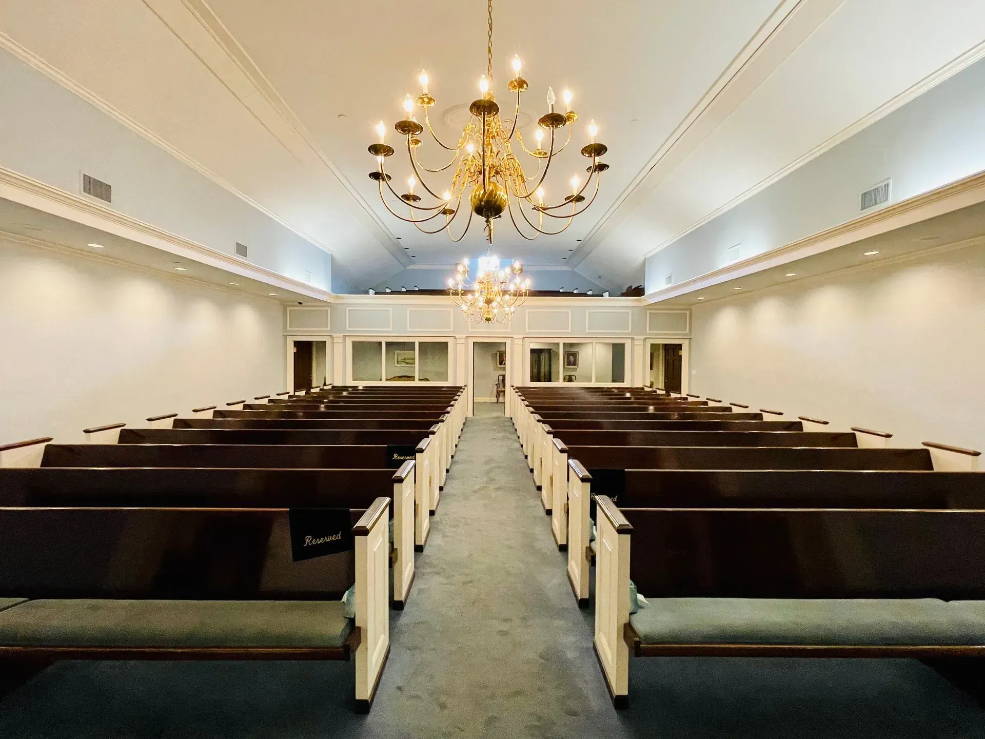 Empty church sanctuary with dark wooden pews, white walls, and chandeliers hanging from the ceiling