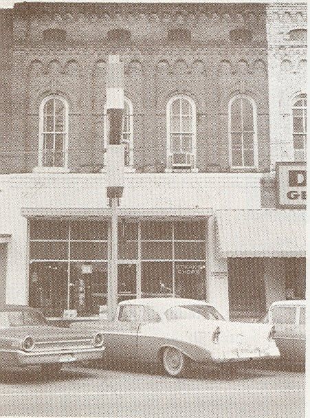 Vintage storefront with two parked cars on a street, including a white convertible in front.