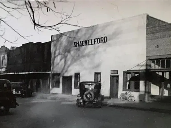 Black-and-white street view of a SHAKLEE FORD dealership with parked vintage cars.