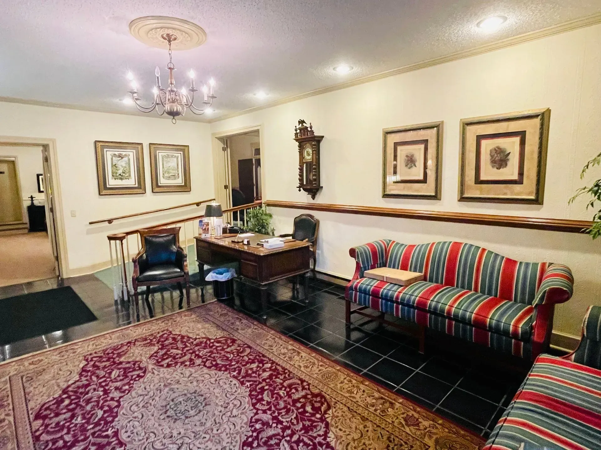 Decorated sitting room with patterned rug, striped sofa, desk, framed art, and chandelier lighting