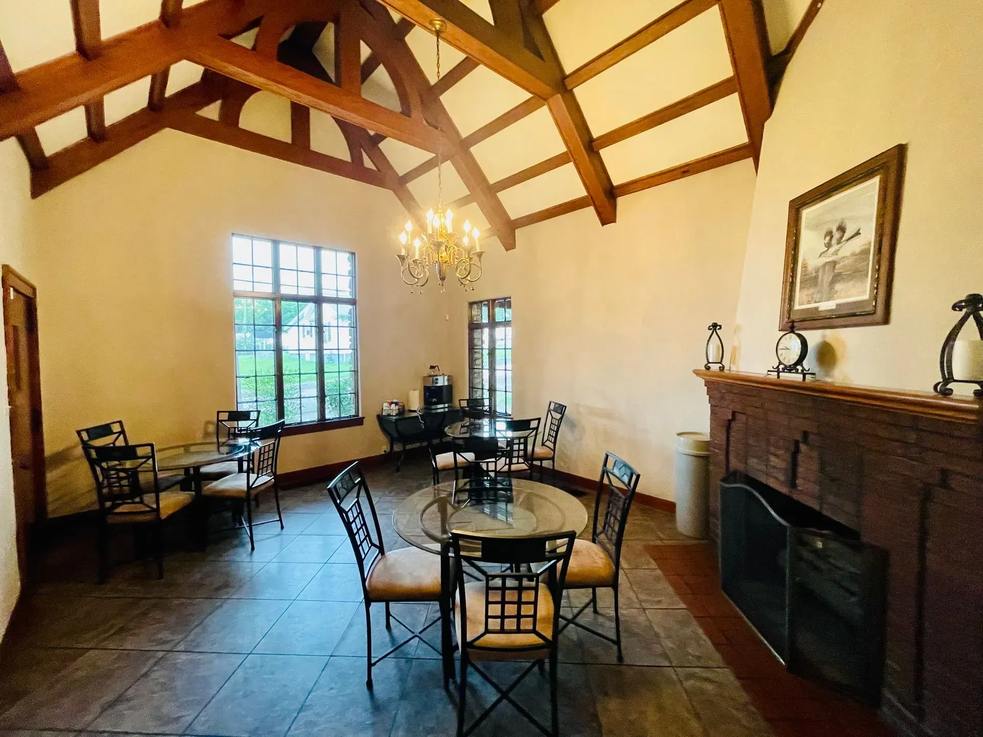 Elegant dining room with high wood beams, chandelier, brick fireplace, and round tables by large windows