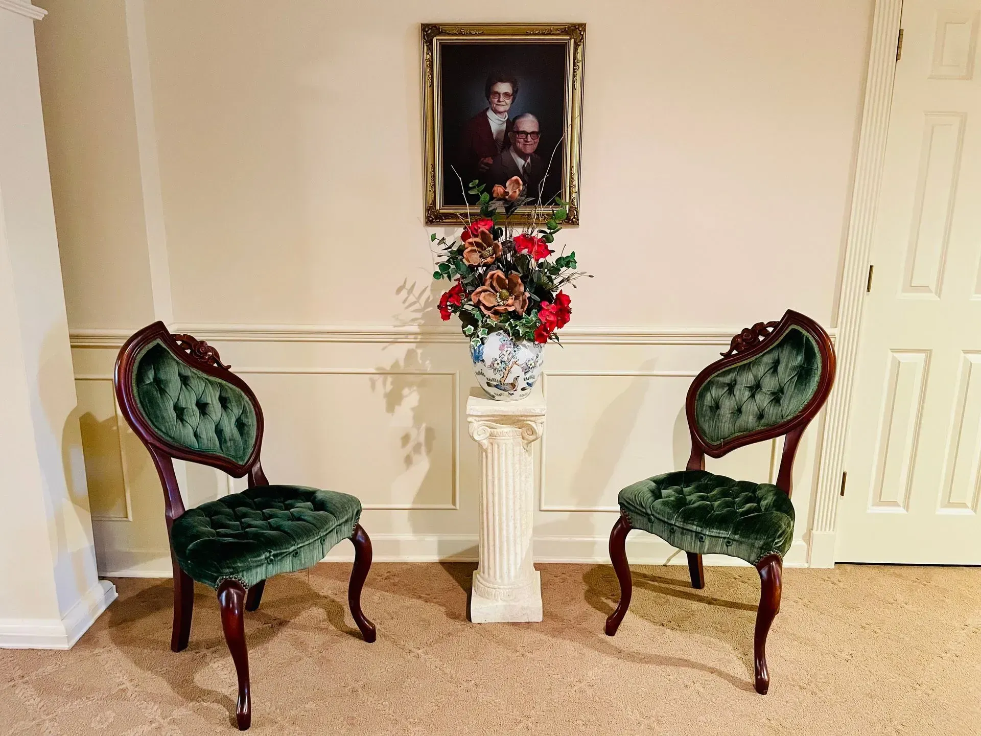 Two green velvet chairs flank a white pedestal with red flowers beneath a framed portrait.
