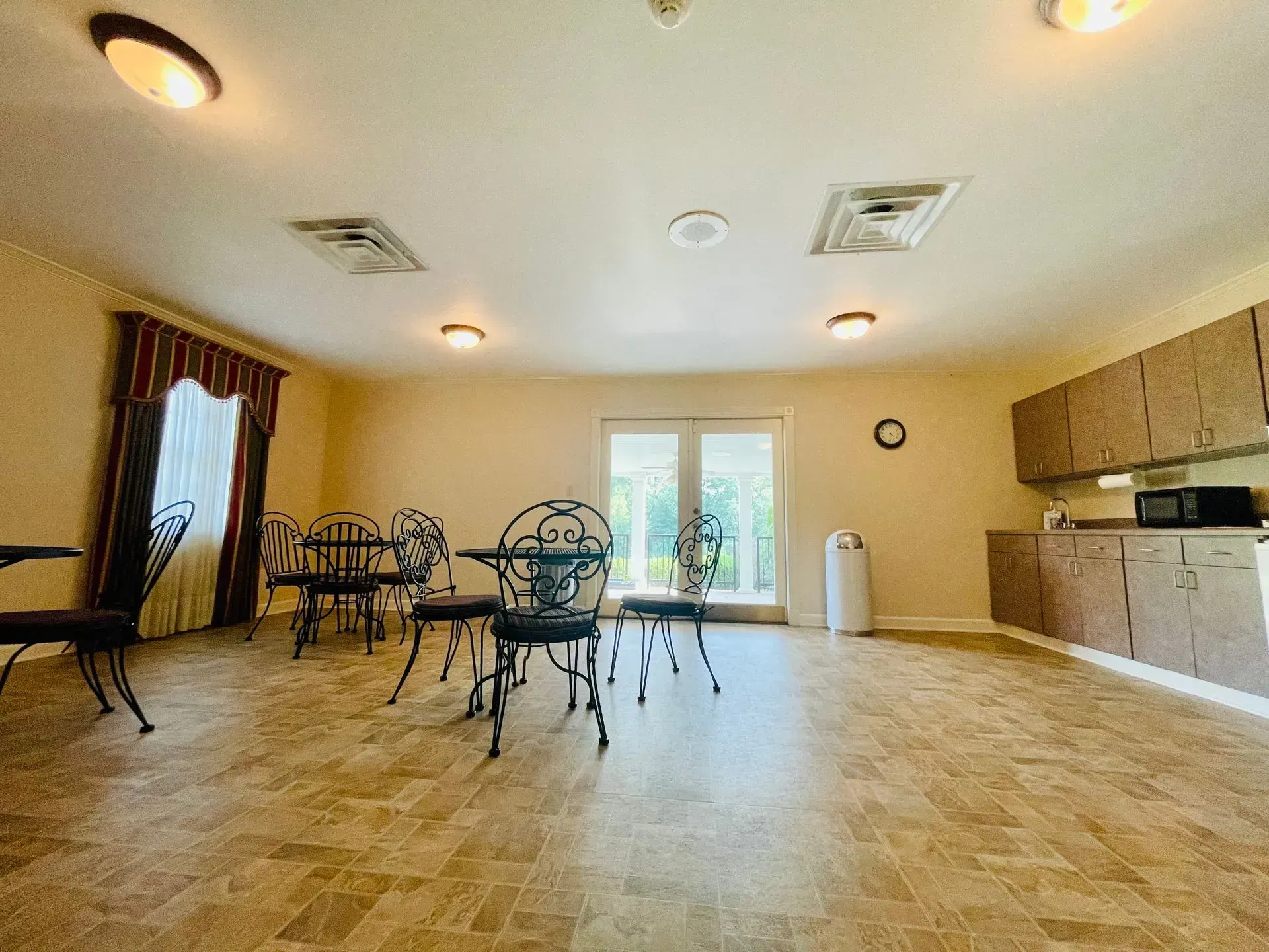Empty community room with tiled floor, beige walls, tables and chairs, cabinets, and a windowed door.