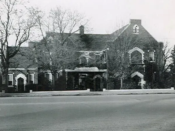 Black-and-white photo of a large brick building with arched windows and a covered entrance beside a road