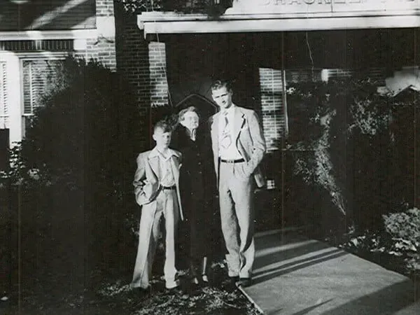 Two people standing on a front porch beside a house, one in a hat, with strong sunlight and shadows