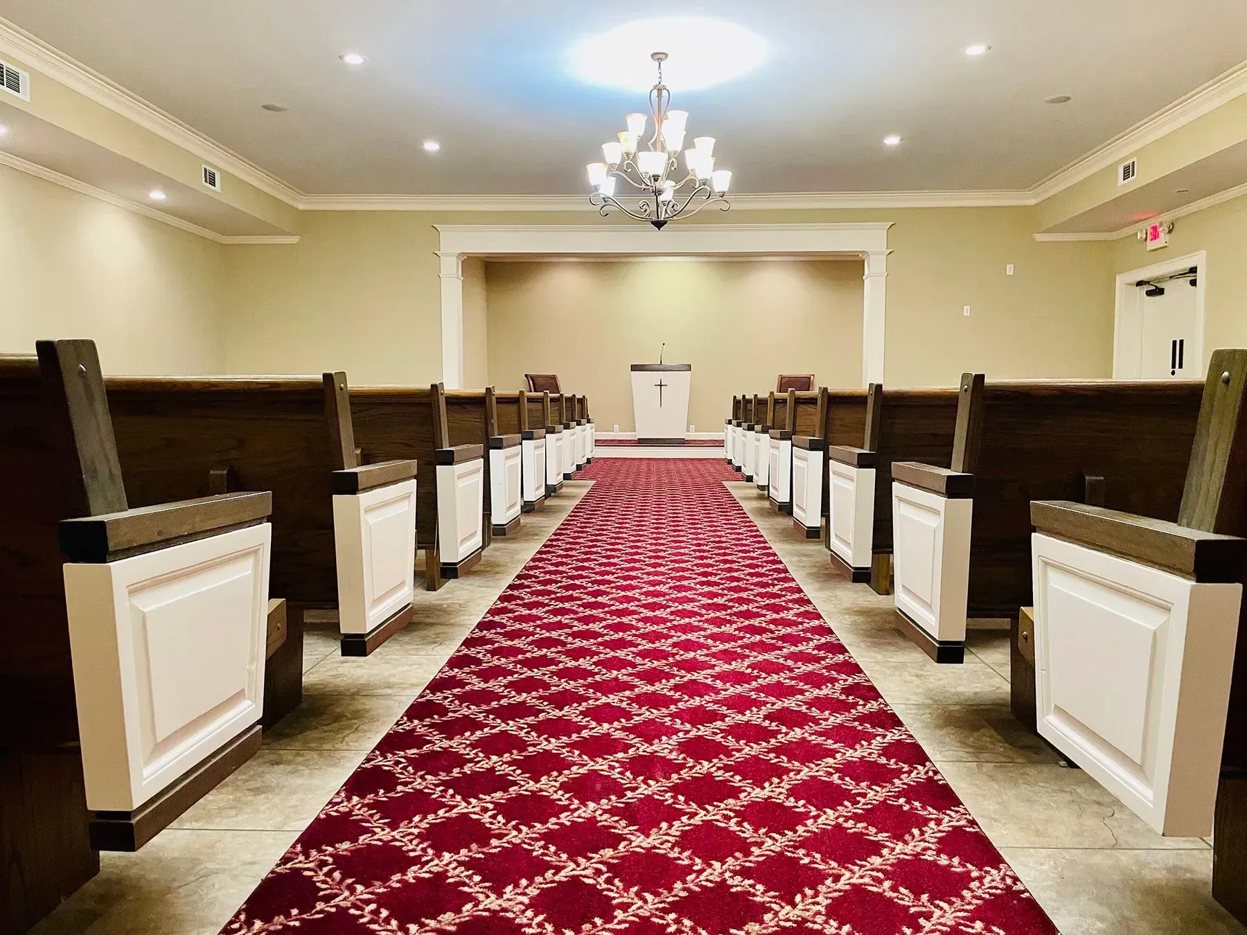 Interior of a small chapel with wooden pews, red carpet, and a chandelier leading to an altar.