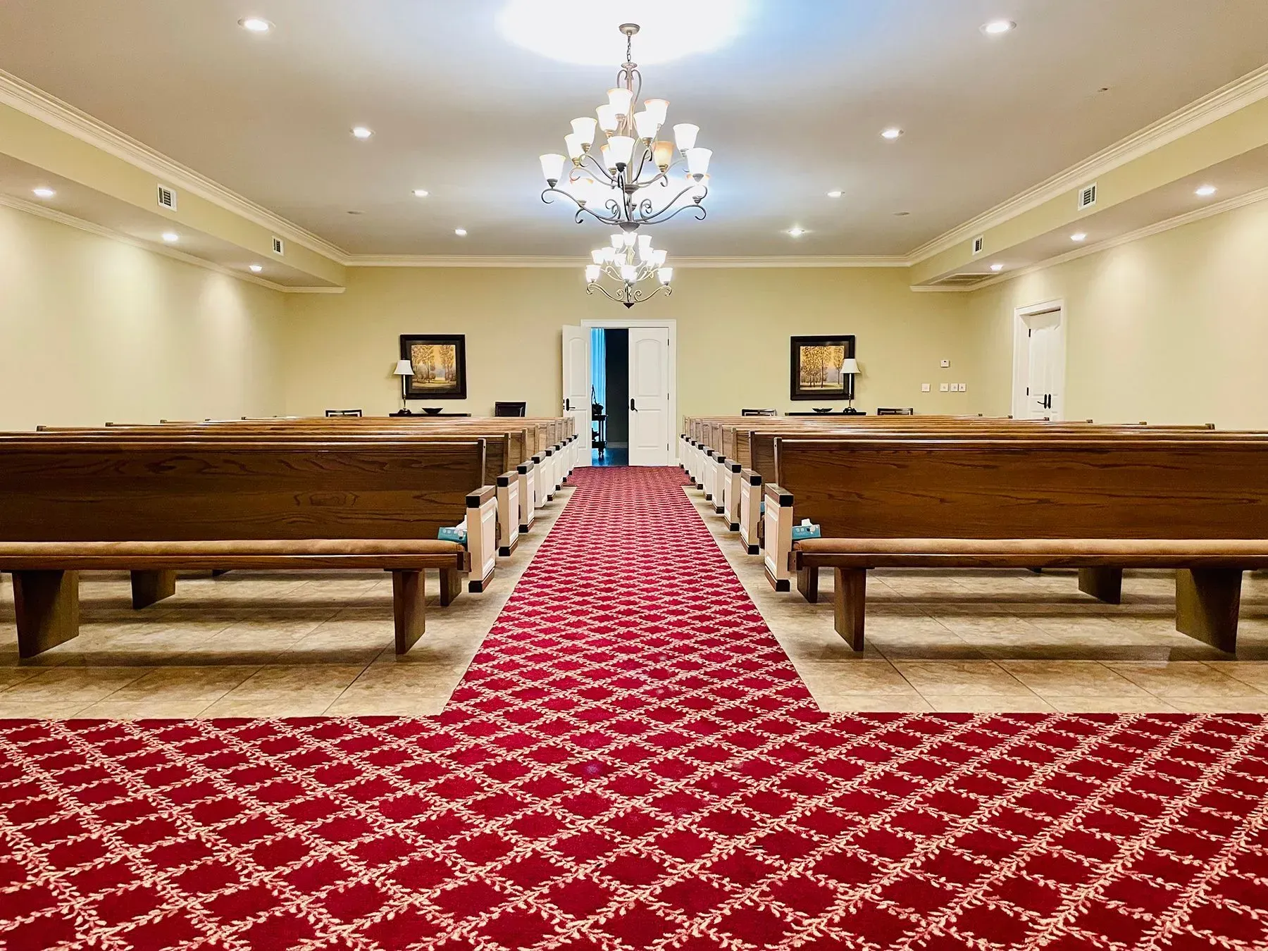 Symmetrical chapel aisle with red carpet, wooden pews, and a chandelier under soft ceiling lights