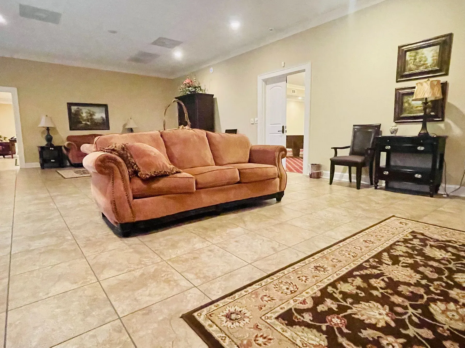 Spacious living room with tan sofa, tiled floor, patterned rug, and wall shelves.