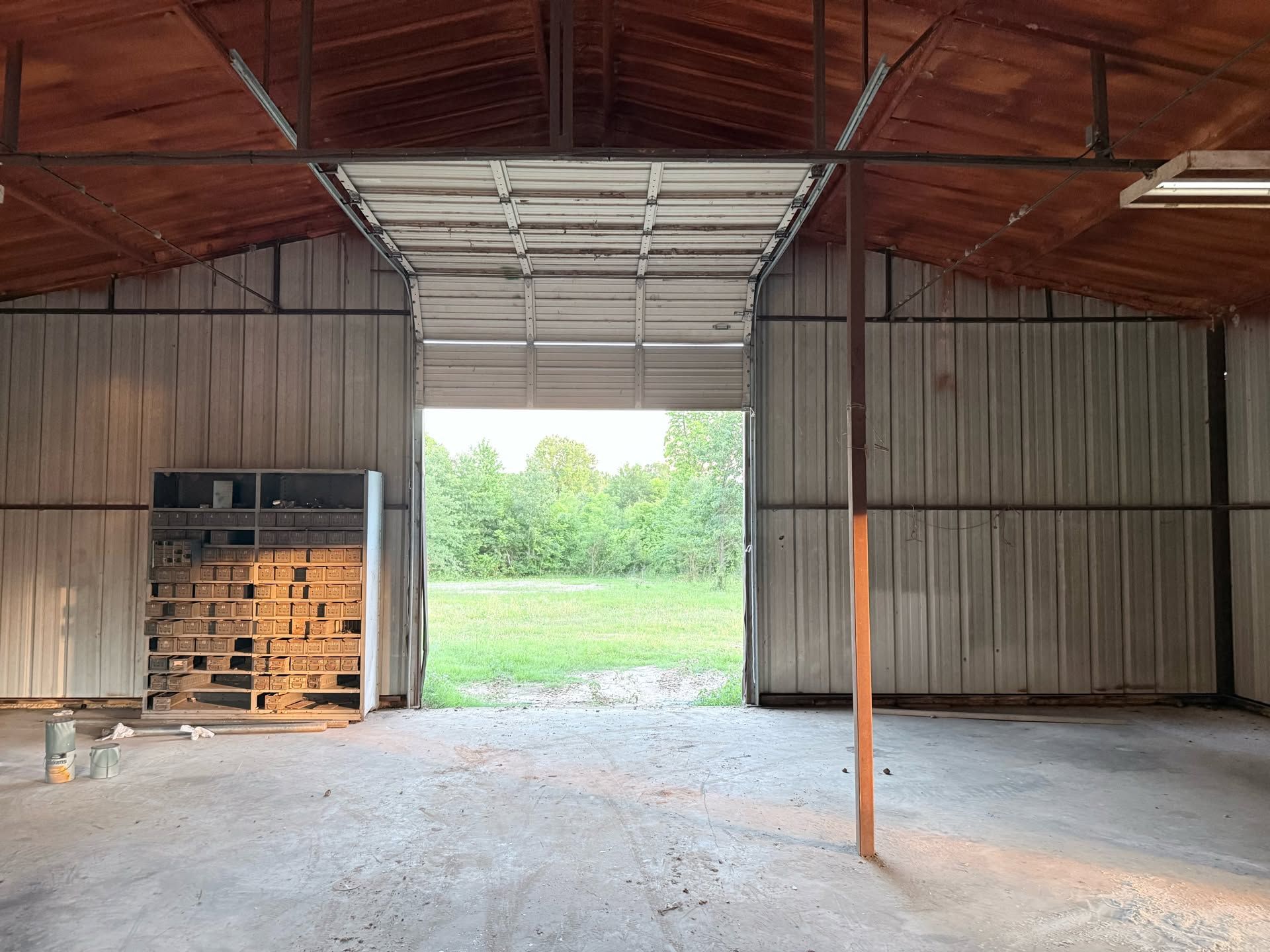 Empty metal warehouse with open roll-up door, concrete floor, stacked pallets, and grassy field outside