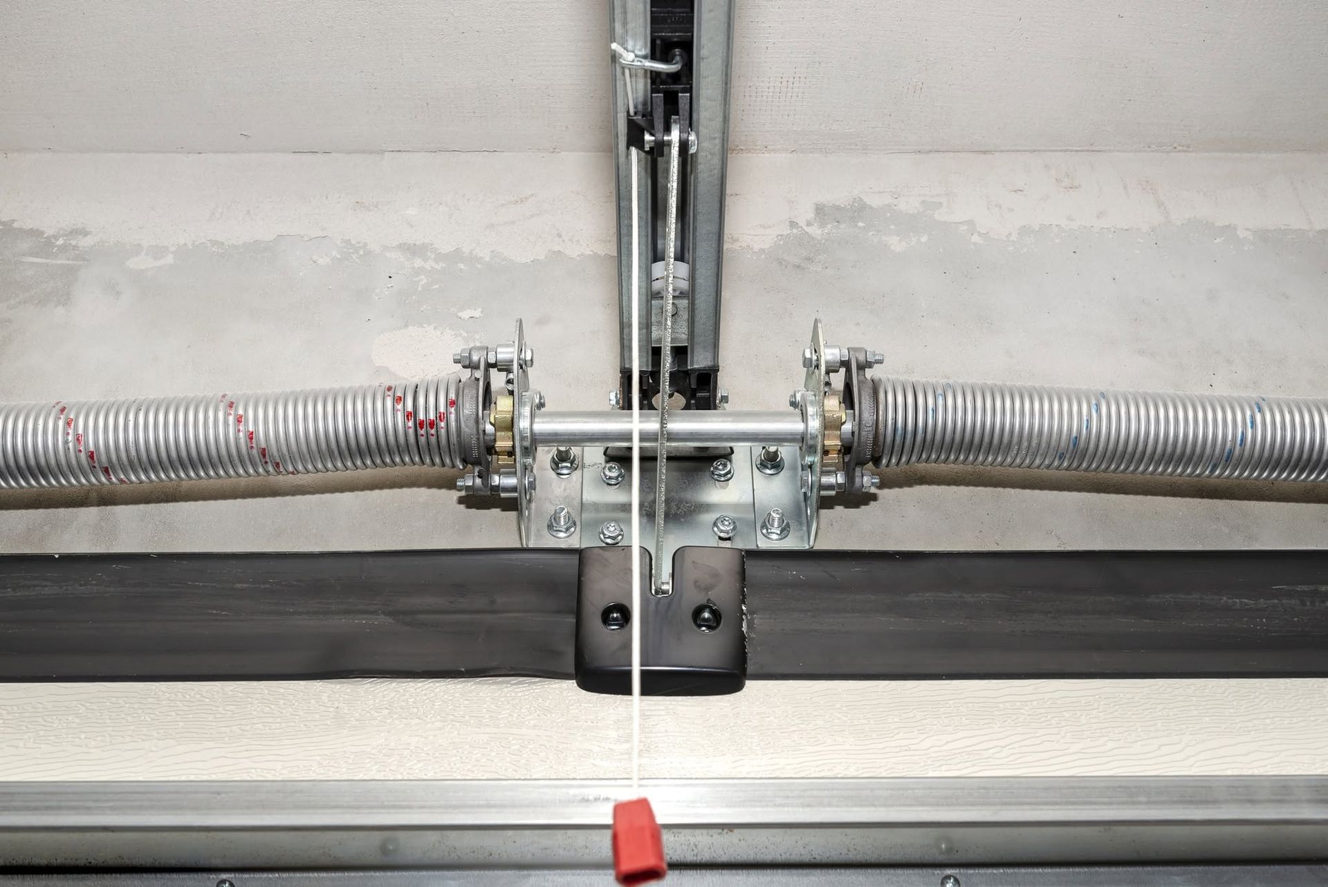 Close-up of a metal garage door opener mechanism with springs and rails against a gray wall.