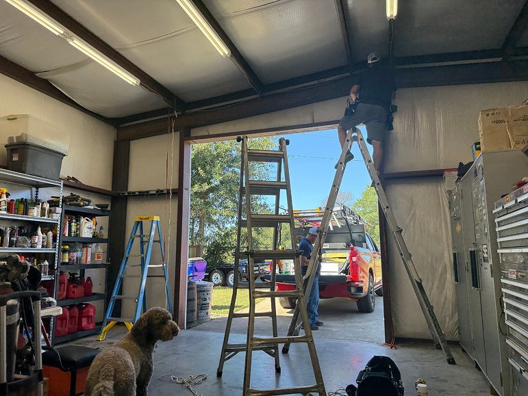 Worker on a ladder in a garage, holding a ceiling panel while a dog watches from below.