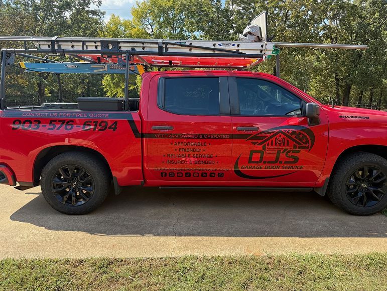 Red pickup truck with ladder rack and black wheels parked beside trees on a roadside.