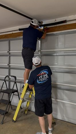 Two workers install a garage door opener, one on a ladder and one standing below.