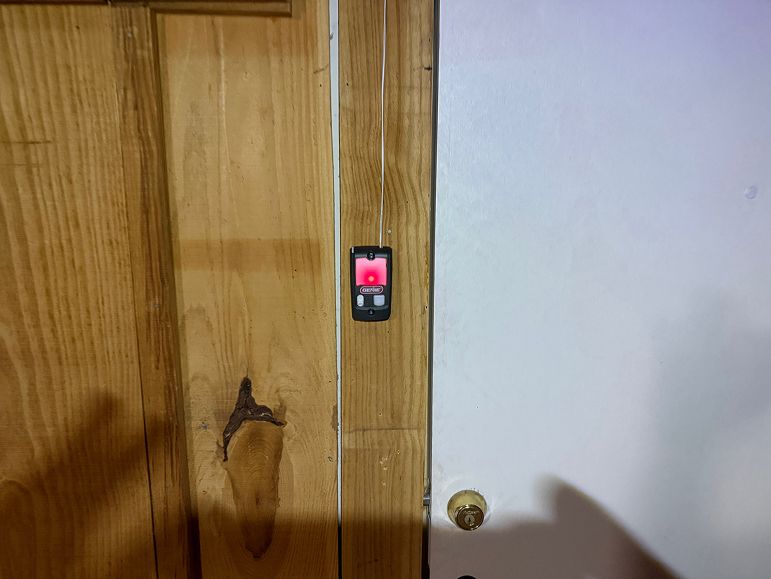Wooden doorframe with a small red illuminated switch beside a white wall and brass doorknob.