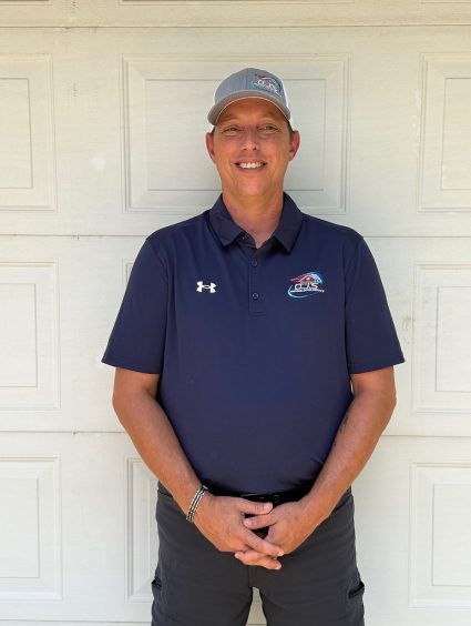 Man standing against a white door, wearing a navy polo, dark pants, and a cap, smiling at the camera