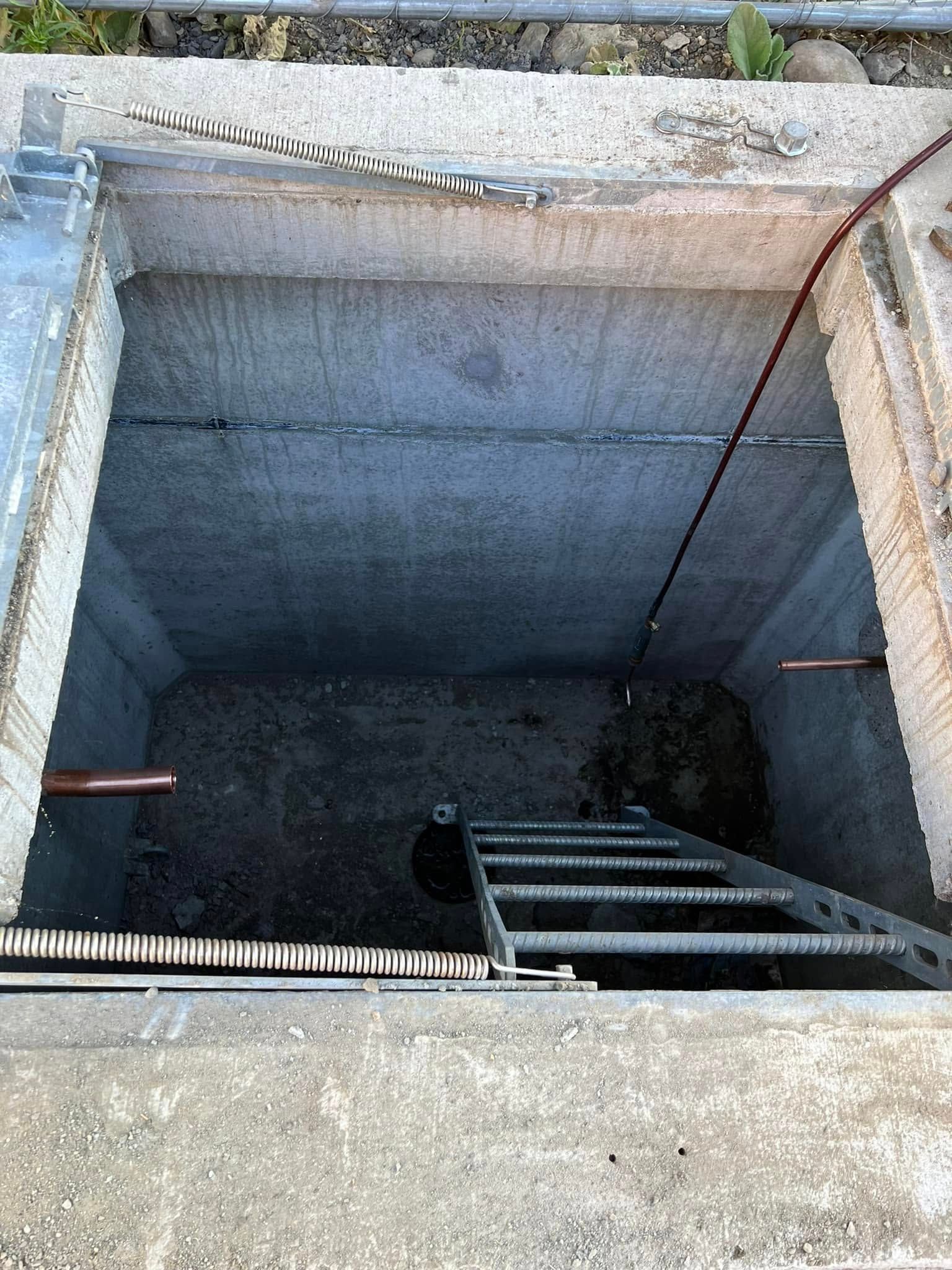An open concrete utility vault showing a descending metal ladder and protruding copper pipes against gray walls.