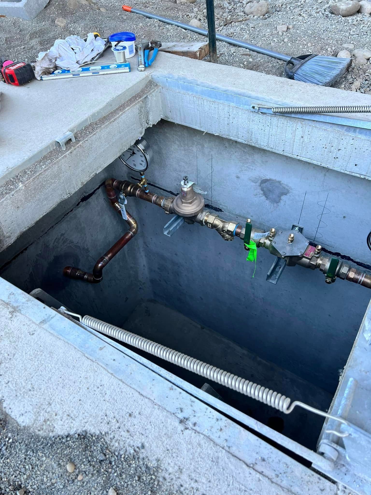 A rectangular concrete utility pit containing copper plumbing pipes and a water meter assembly.