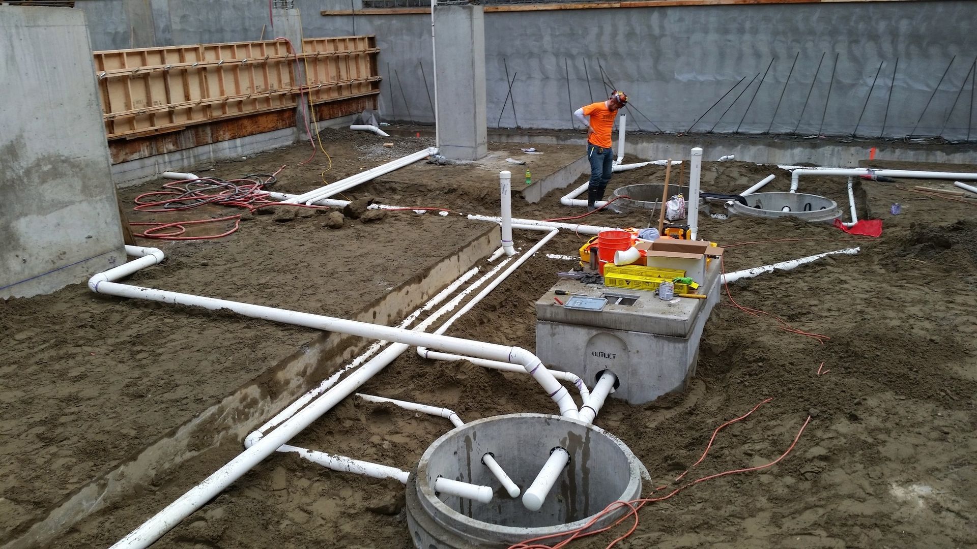 A construction worker stands in a dirt basement foundation installing white PVC plumbing pipes and utility conduits.