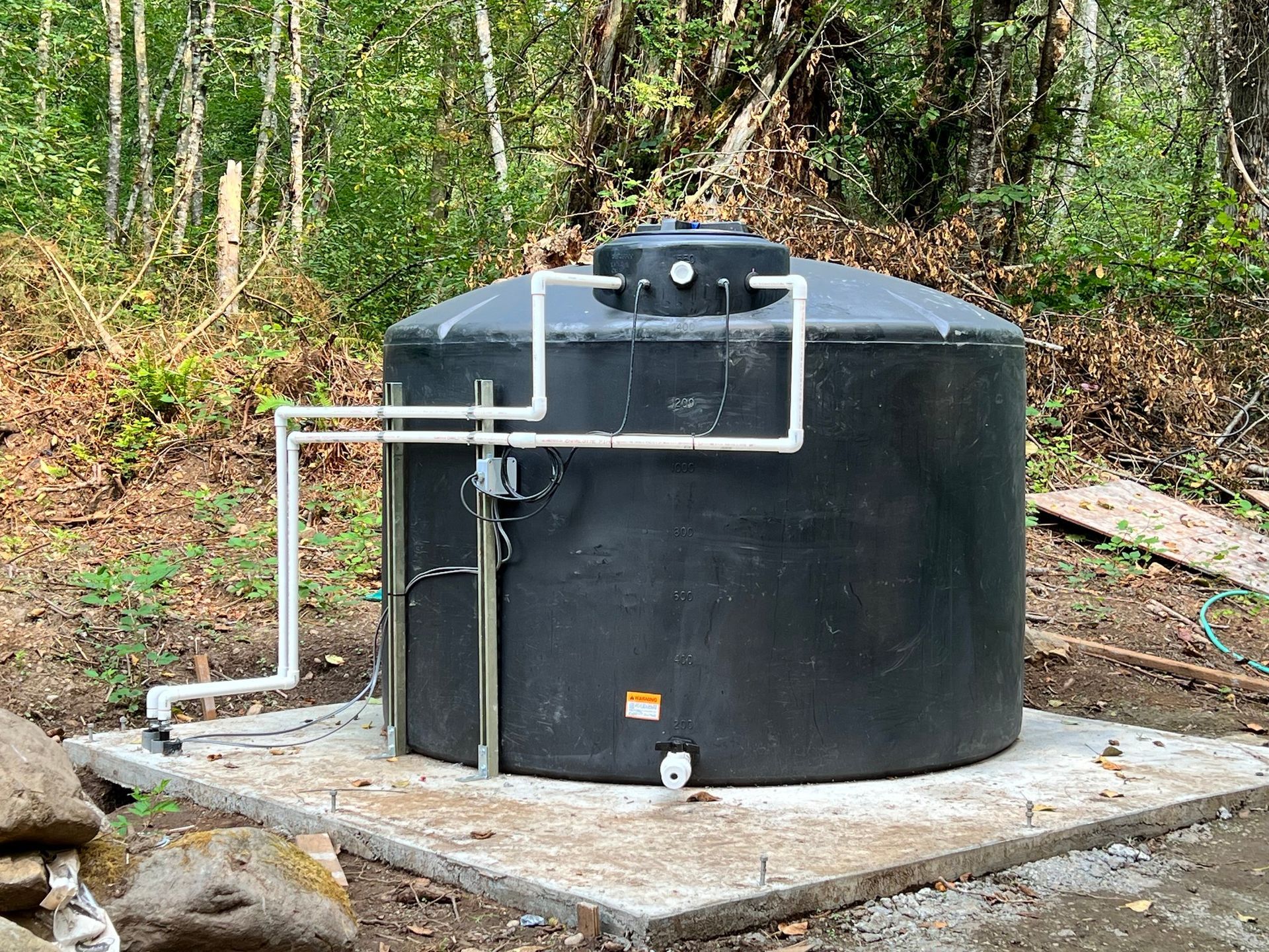 A large, black circular water storage tank sits on a concrete pad in a wooded area, connected to external white piping.