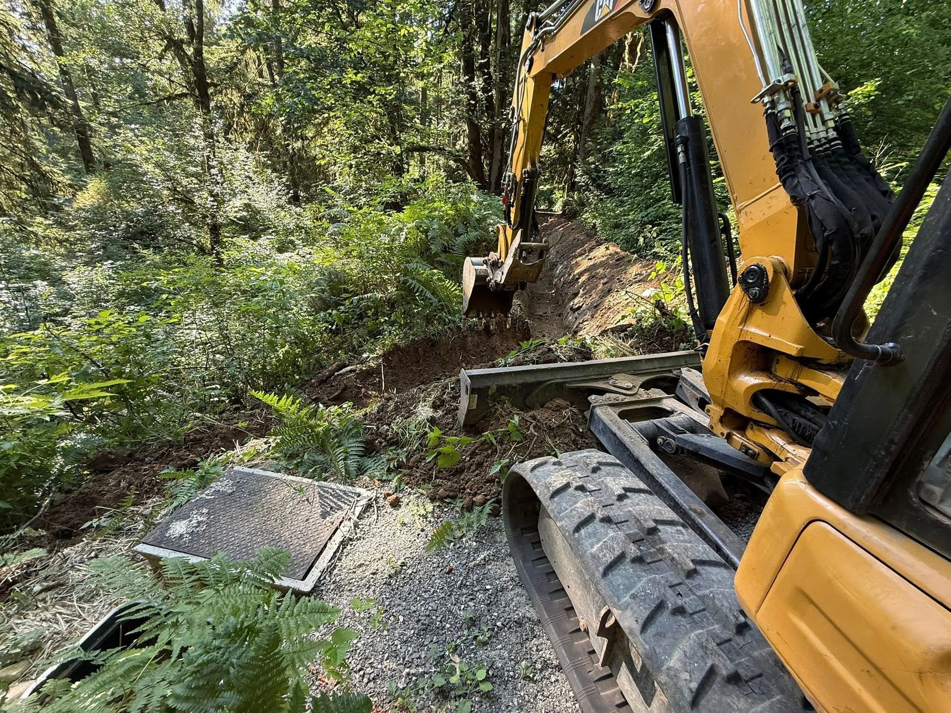 A yellow excavator parked on a gravel path in a wooded area, with a small rectangular utility cover in the foreground.