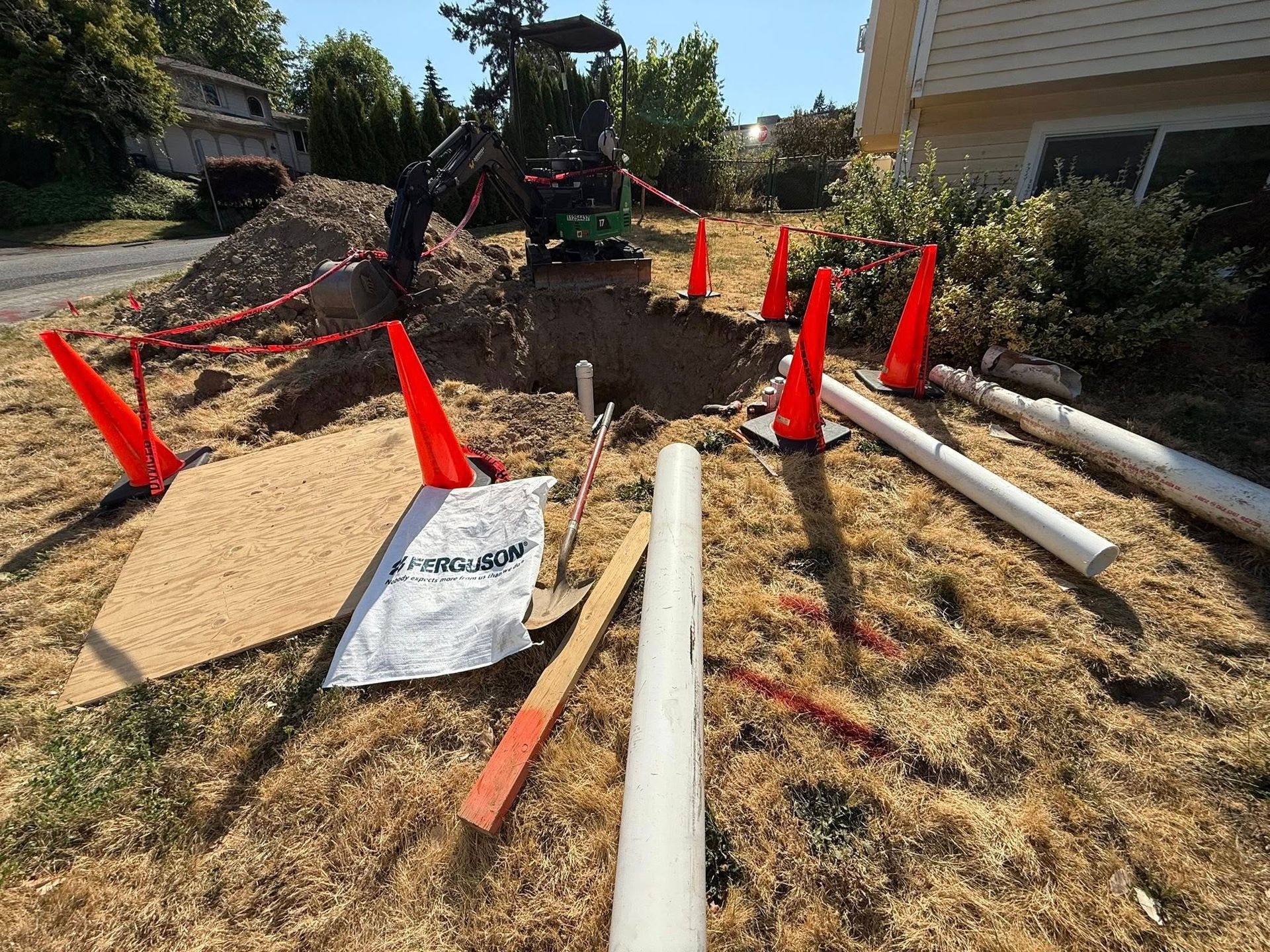 A mini excavator digs a hole in a yard near a house, marked by orange safety cones and sections of white PVC pipe.